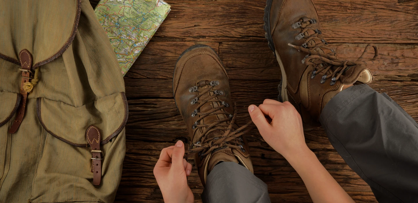 Person tying brown hiking boots next to a green backpack and a map on a wooden floor.